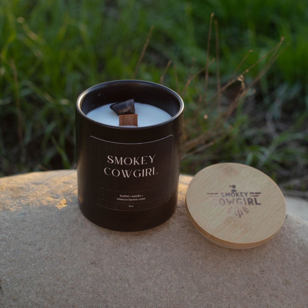 Smokey Cowgirl candle in a black container with wooden lid on a stone surface with grass in the background