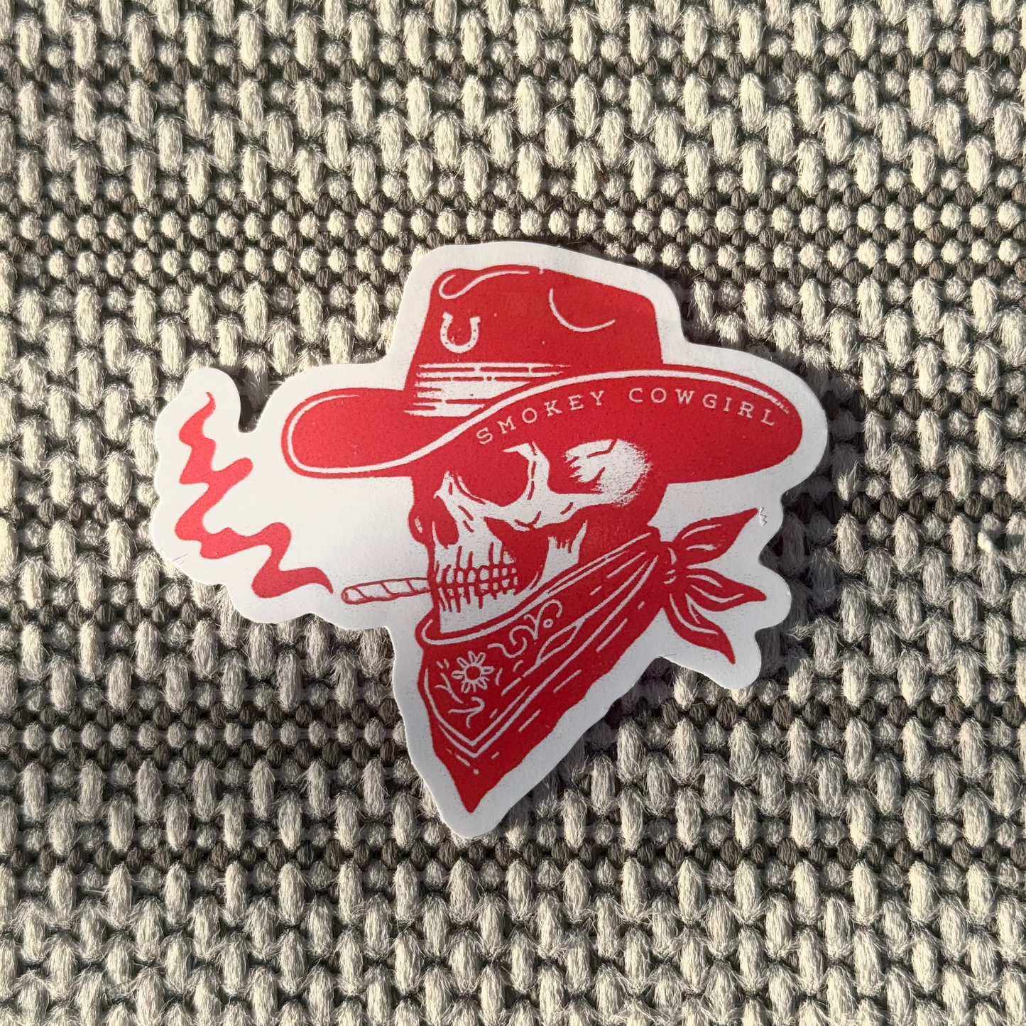 Red 'Smokin' Cowgirl' sticker with skull design on a textured fabric background