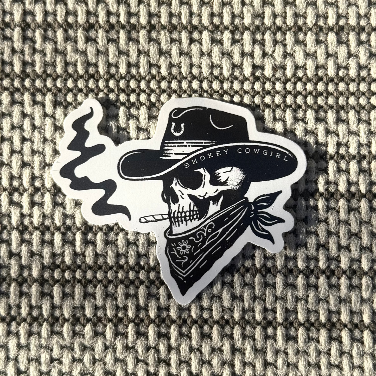 Sticker of a skull wearing a cowboy hat and bandana with 'Smookey Cowgirl' text on a textured fabric background.